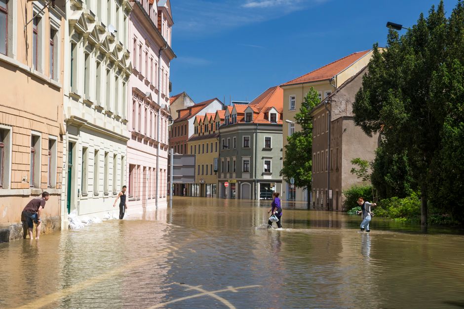 Strada europea allagata con persone che camminano nell'acqua alta, simbolo dell'impatto del cambiamento climatico sull'edilizia scolastica e le aule.