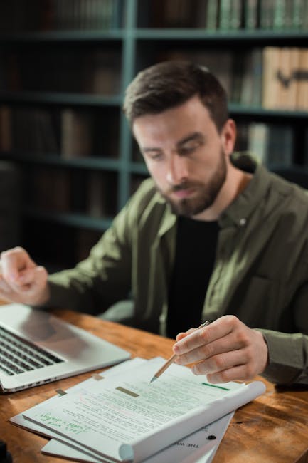 Studioso analizza capolavoro letterario con laptop e documenti in biblioteca, concentrato sulla profondità dell'opera e il suo impatto culturale.