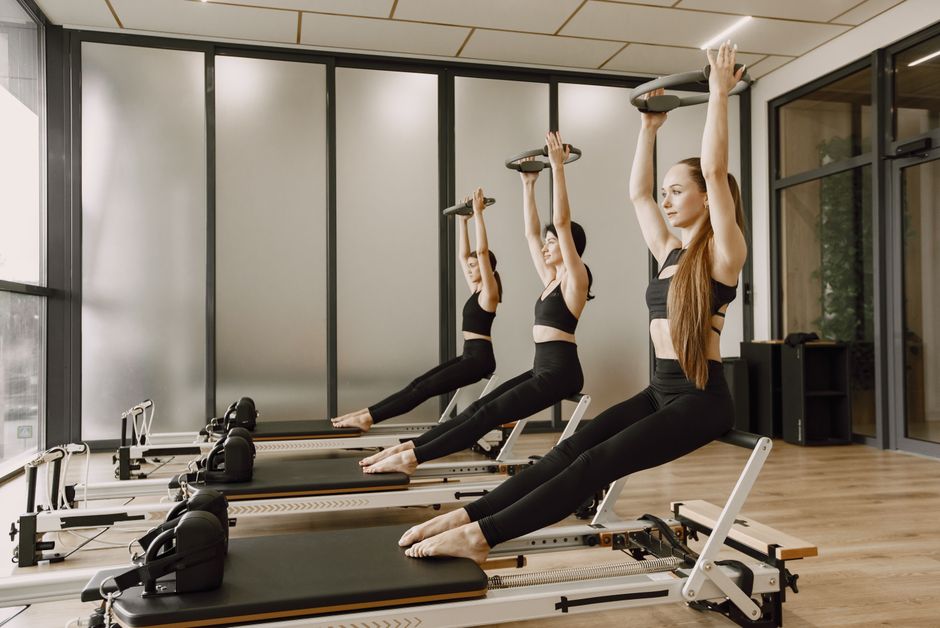 Tre donne in abiti sportivi neri eseguono esercizi di pilates su reformer con anelli di resistenza sollevati sopra la testa in una moderna palestra