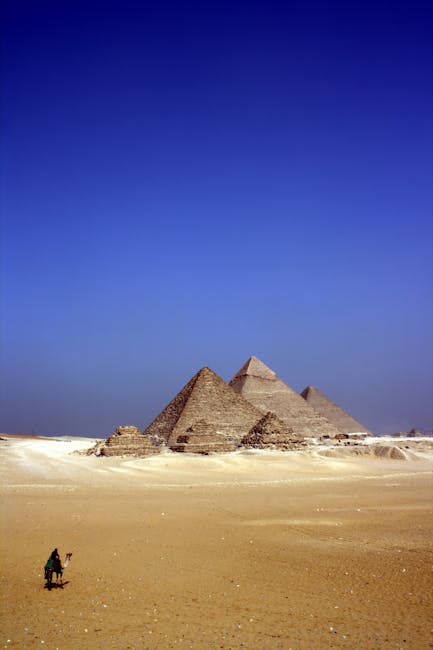 Le maestose piramidi di Giza, testimonianza millenaria dell'Antico Egitto, con un cammello che attraversa il deserto dorato sotto un cielo azzurro intenso.